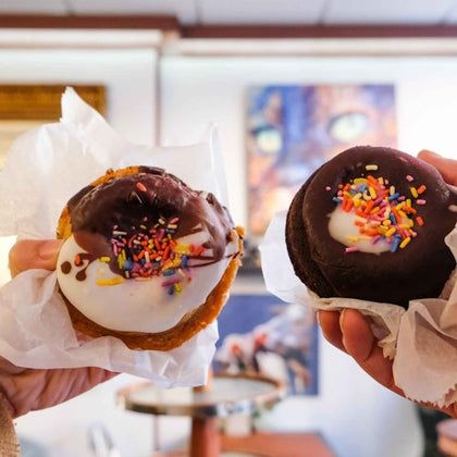 Two hands hold up pastries covered in white and chocolate icing with colorful sprinkles in a cafe setting.
