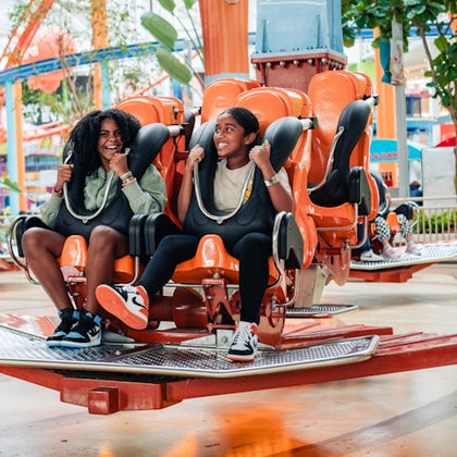 Two people smiling and seated on an orange amusement ride, surrounded by a vibrant indoor park setting.