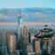 A helicopter flies over Manhattan with One World Trade Center and surrounding skyscrapers in the background under a partly cloudy sky.