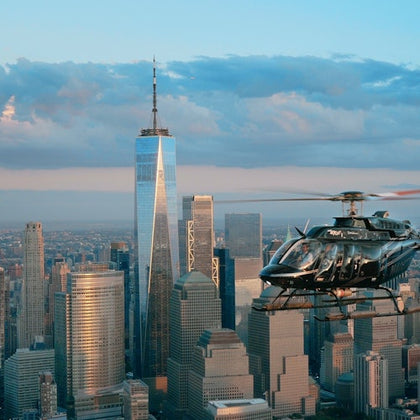 A helicopter flies over Manhattan with One World Trade Center and surrounding skyscrapers in the background under a partly cloudy sky.