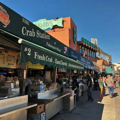 People walking past a row of outdoor seafood vendors at Fisherman's Wharf, including "The Crab Station." Signs advertise fresh crab and clam chowder.