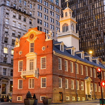 A historic red-brick building with white trim and a clock tower, surrounded by modern skyscrapers, under evening light.