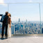 Two people standing on a high observation deck with a city skyline in the background, taking a selfie.