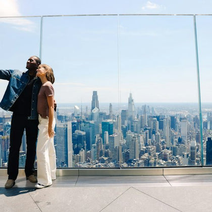 Two people standing on a high observation deck with a city skyline in the background, taking a selfie.