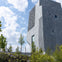 A modern concrete building with a textured facade stands amidst landscaped greenery under a partly cloudy sky.
