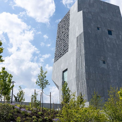 A modern concrete building with a textured facade stands amidst landscaped greenery under a partly cloudy sky.