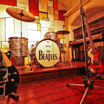 A vintage drum set with "The Beatles" logo, guitars, and a piano on a wooden stage with colorful wall panels.