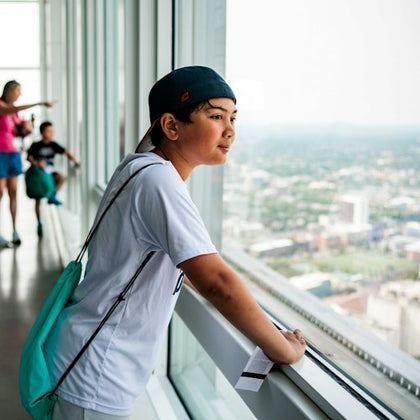 A boy in a white shirt and baseball cap looks out a large glass window in a high-rise building, with a cityscape in the background.