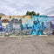 A colorful mural featuring a raccoon in a blue hoodie sitting amidst an urban scene with flowers and buildings under a cloudy sky.