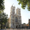 A large Gothic-style cathedral with two tall towers, surrounded by trees and a clear sky, with some people walking nearby.