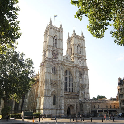 A large Gothic-style cathedral with two tall towers, surrounded by trees and a clear sky, with some people walking nearby.