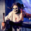 A person in a horned skull mask and bloody white robe holds a red axe in a dimly lit room.