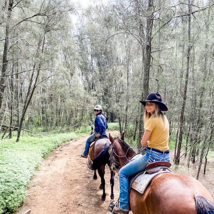 Two people riding horses on a dirt path in a forested area, with trees and greenery surrounding them.