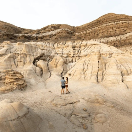 Two people stand on a rocky, eroded landscape with striated formations, under a light sky.
