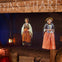 Two women dressed in historical attire stand in a dimly lit scene resembling an old wharf, with barrels and lanterns nearby.