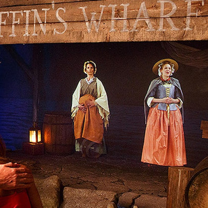 Two women dressed in historical attire stand in a dimly lit scene resembling an old wharf, with barrels and lanterns nearby.