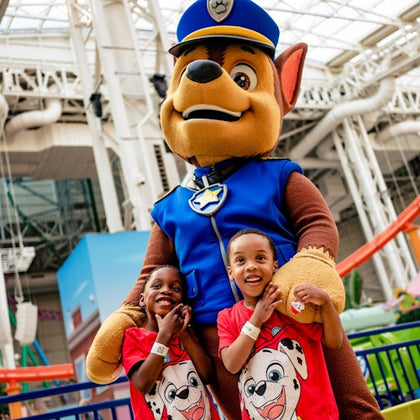 Two children smiling and posing with a person in a large dog costume wearing a police uniform in an indoor amusement setting.