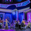 A colorful stage play scene with actors in formal attire seated and standing in a lavishly decorated room.