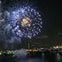 A nighttime scene of fireworks bursting over a city skyline with illuminated buildings and a body of water in the foreground.