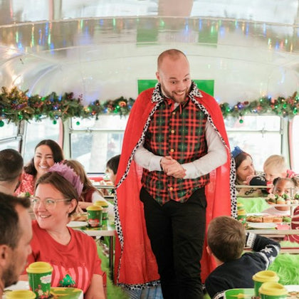 A festive scene inside a decorated bus with people enjoying food and a man in a red cape interacting with them.