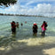 People wearing life vests, standing in shallow water near a beach, with a city skyline in the background under a partly cloudy sky.