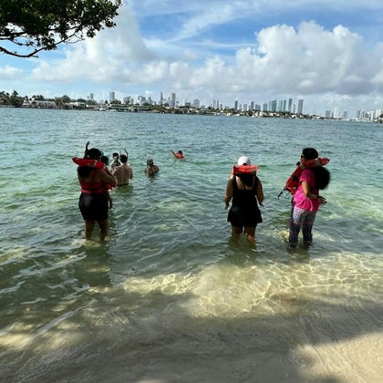People wearing life vests, standing in shallow water near a beach, with a city skyline in the background under a partly cloudy sky.