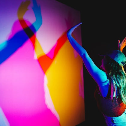 Person in a cap interacting with a multicolored light installation, casting overlapping shadows on the wall.