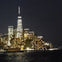 A city skyline at night with illuminated skyscrapers reflected on water, dominated by a tall, lit tower.