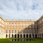 Symmetrical courtyard with a round central fountain, surrounded by a red-brick building with arched windows under a partly cloudy sky.