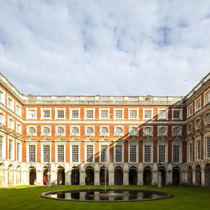 Symmetrical courtyard with a round central fountain, surrounded by a red-brick building with arched windows under a partly cloudy sky.