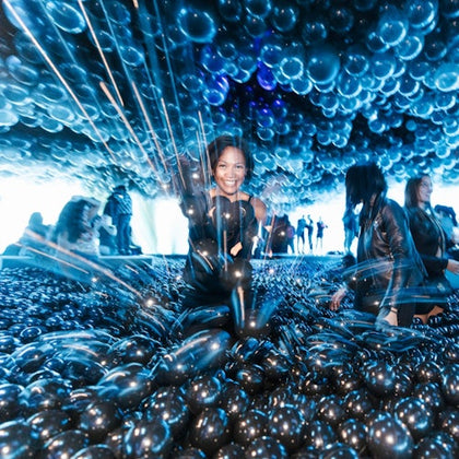 A person smiling in a room filled with black balloons, with others in the background under blue lighting.