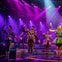 Performers on stage in colorful costumes under purple and blue lighting, with musicians visible in the background.