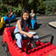 Children driving colorful, toy-like cars in an outdoor amusement park with lush greenery in the background.