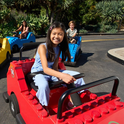 Children driving colorful, toy-like cars in an outdoor amusement park with lush greenery in the background.