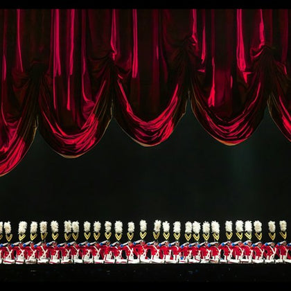 A line of performers dressed as toy soldiers in red uniforms and white hats stands under red stage curtains.