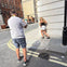 A man photographing a woman posing against the corner of a building on a city street during daylight.