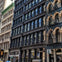 A city street with multi-story buildings featuring ornate facades and fire escapes on a sunny day.