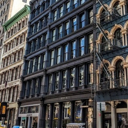 A city street with multi-story buildings featuring ornate facades and fire escapes on a sunny day.