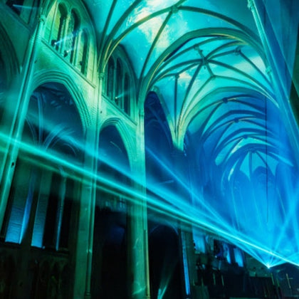Interior of a cathedral illuminated with blue lights and laser beams, showing arched ceilings and ornate architecture.