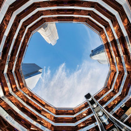 Upward view through an octagonal structure with copper-colored lining, revealing skyscrapers and blue sky with scattered clouds.