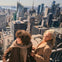 Two people smiling on a rooftop overlooking a city skyline with tall buildings under a clear sky.