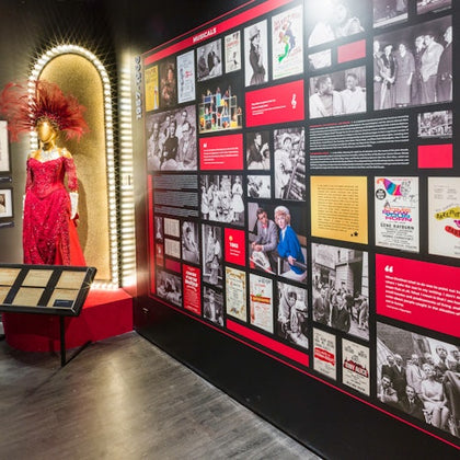 Exhibit with a red sequined dress on a mannequin and a well-lit wall display featuring black-and-white photos and historical documents.