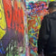 A person wearing a black jacket and cap standing in front of a wall covered with colorful graffiti.