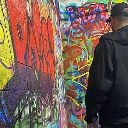 A person wearing a black jacket and cap standing in front of a wall covered with colorful graffiti.