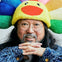 Person with round glasses, long hair, and a colorful plush hat shaped like a sunflower, smiling and resting hands under chin.