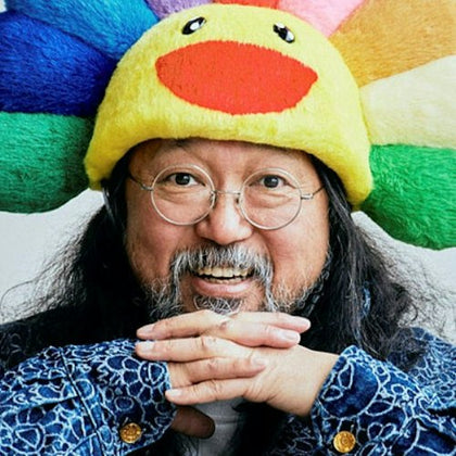 Person with round glasses, long hair, and a colorful plush hat shaped like a sunflower, smiling and resting hands under chin.