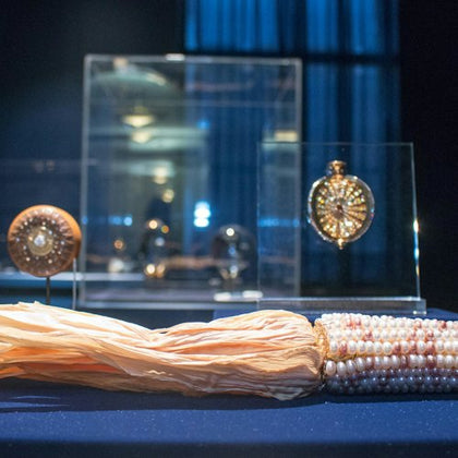 A collection of ornate jewelry displayed in glass cases, with a pearl and tassel necklace in the foreground on a blue surface.