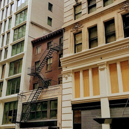 A city building facade with fire escapes on a red structure between beige and white buildings.