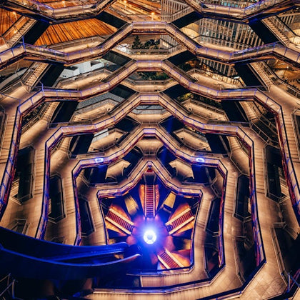 Geometric structure with multiple hexagonal levels, illuminated by purple and yellow lights, seen from an upward perspective.