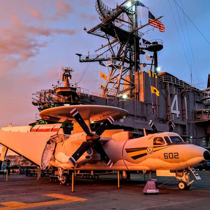 A military aircraft, marked "602," is parked on the deck of a ship with a large structure marked "41" and various flags in the background.